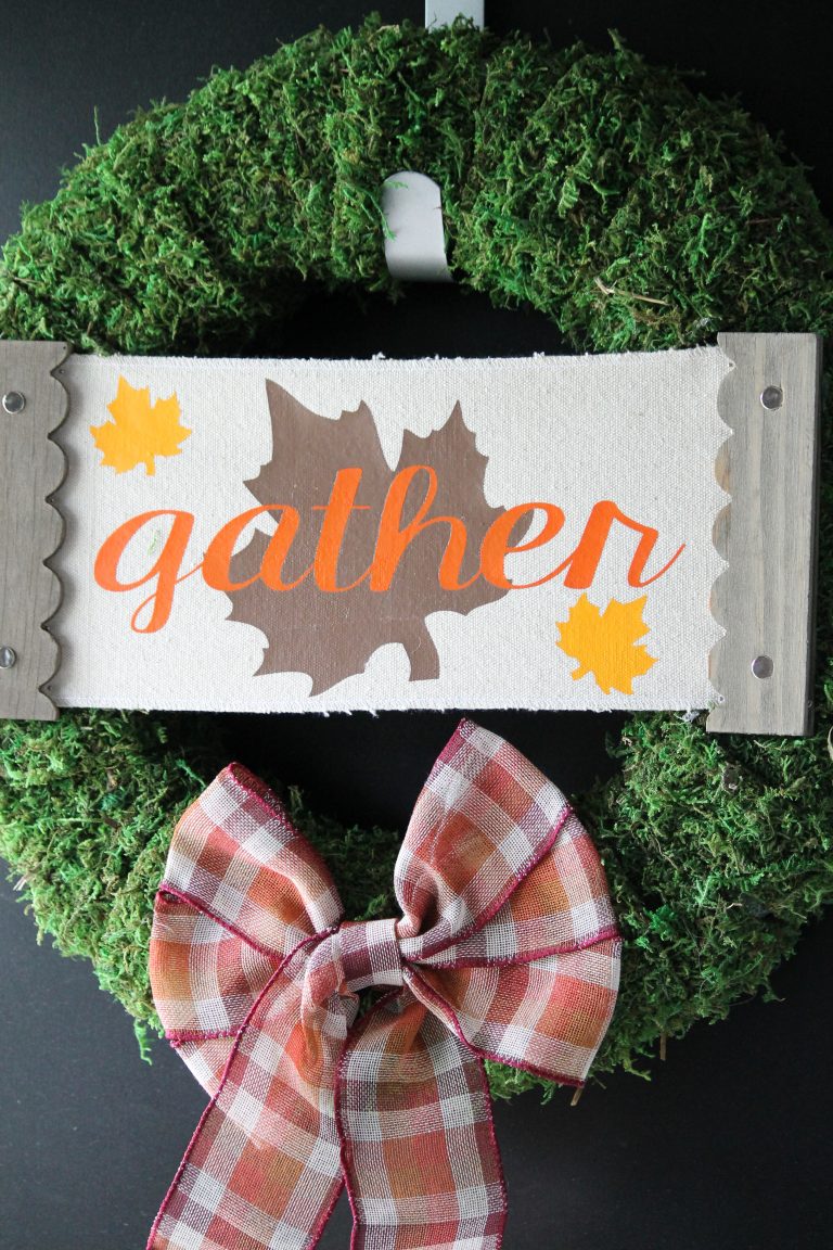 Moss covered fall wreath decorated for fall with a gather sign and a plaid bow