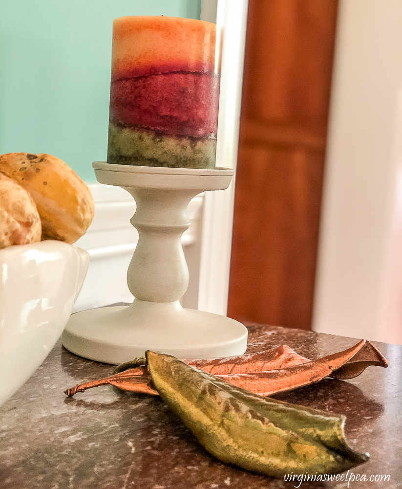 Orange, burgundy, and green fall candle on a white candle holder with bronzed Magnolia leaves