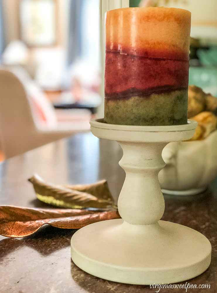 Orange, burgundy, and green fall candle on a white candle holder with bronzed Magnolia leaves