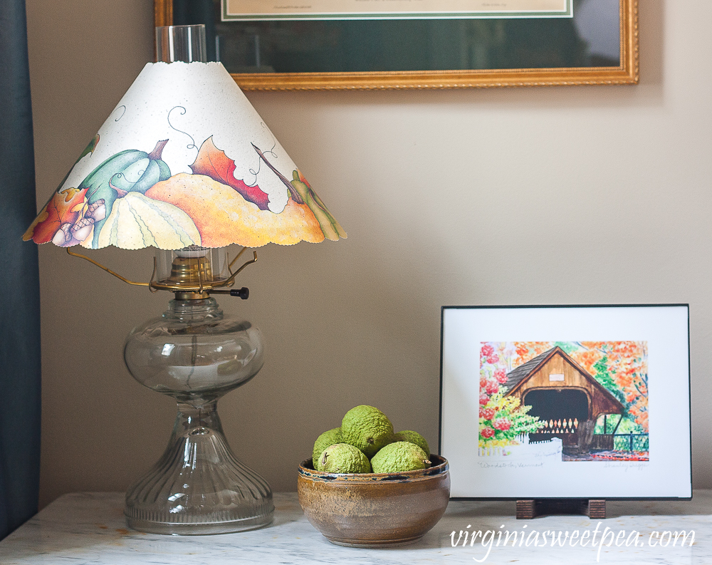 Watercolor of a covered bridge in Woodstock, Vermont with a Walnuts in a North Carolina pottery bowl filled with walnuts and an antique lamp with a paper shades with pumpkins, gourds, and leaves on it.