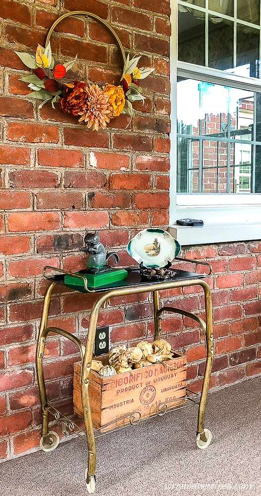 Enclosed porch decorated for fall with a fall hoop wreath, tv cart with a crate of gourds, and a squirrel nutcracker