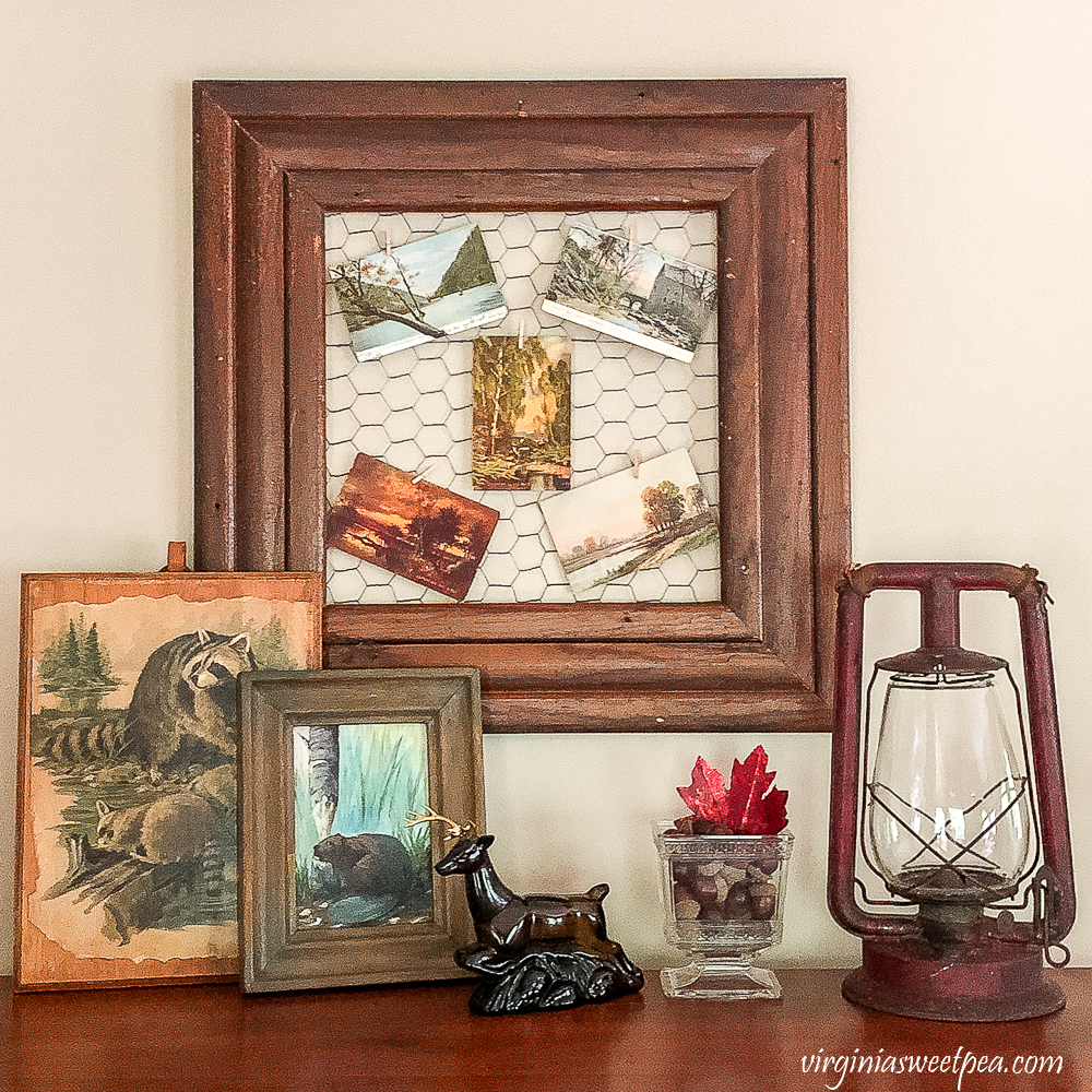 Fall decor with early 1900s postcards, vintage Avon stag cologne bottle with a beaver painting, raccoon decoupaged to a board, vintage red lantern, vintage glass compote filled with squirels and a red leaf.