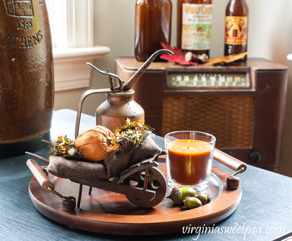 Fall vignette with a Byer's Choice fall decorated wheelbarrow, vintage copper colored oil can, orange candle in a glass holder, acorns with vintage brown bottles in the background