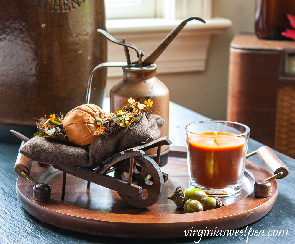 Fall vignette with a Byer's Choice fall decorated wheelbarrow, vintage copper colored oil can, orange candle in a glass holder, acorns