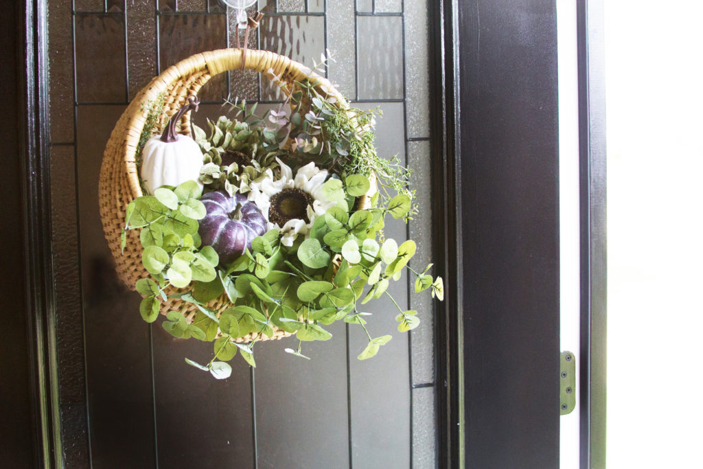 Basket hanging on a front door filled with greenery, a white sunflower, a white pumpkin, and a purple pumpkin