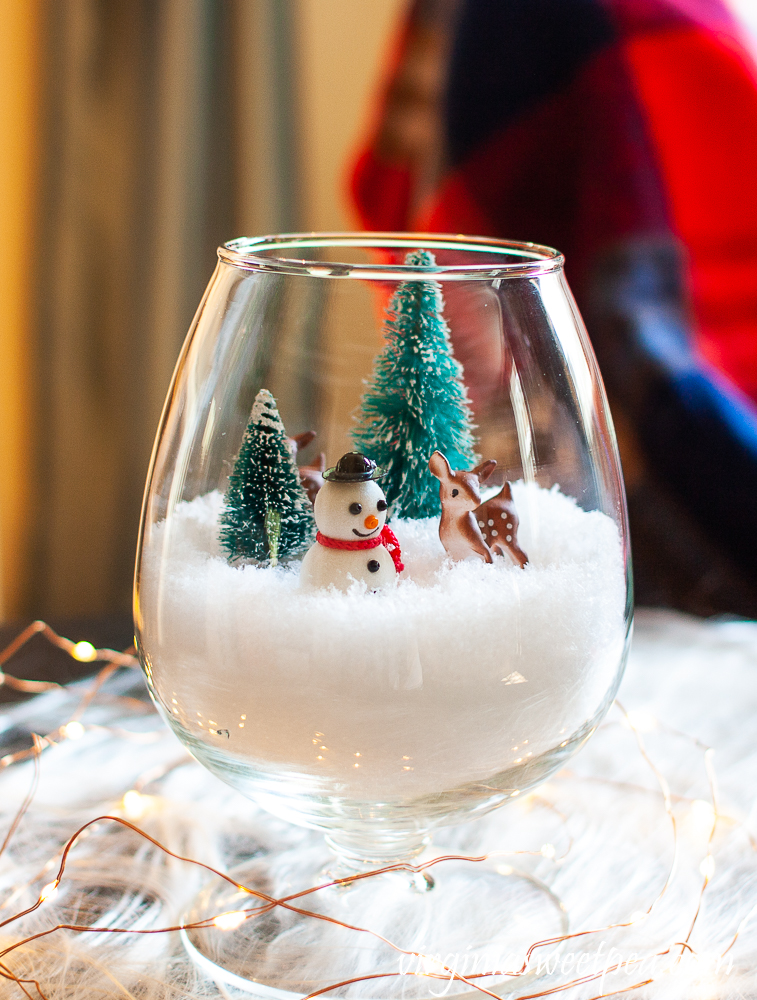 Christmas decoration with a brandy sniffer with faux snow and miniature trees, deer, and a snowman surrounded by twinkle lights.