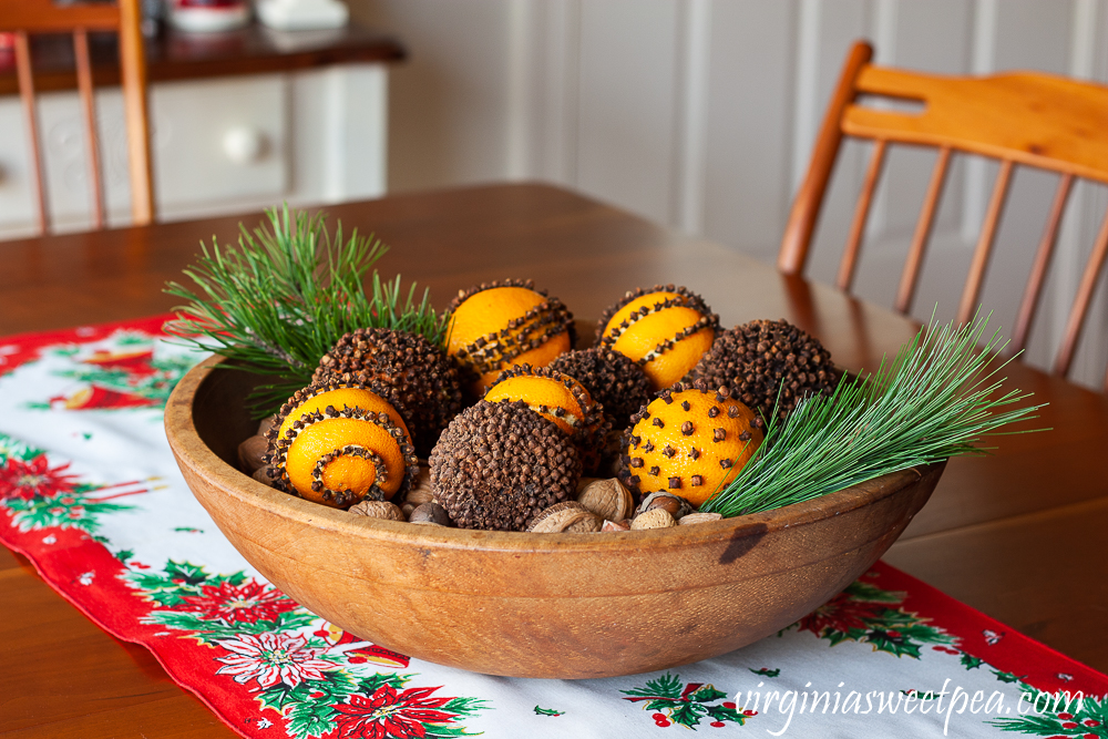 Orange pomanders in a wooden bowl with mixed nuts on the bottom and two sprigs of pine