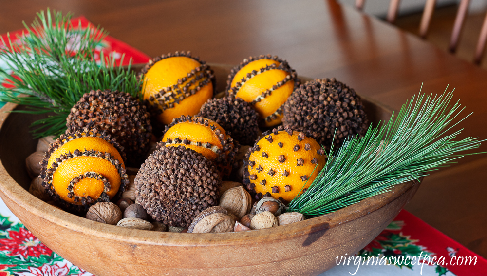 Orange pomanders in a wooden bowl with mixed nuts on the bottom and two sprigs of pine