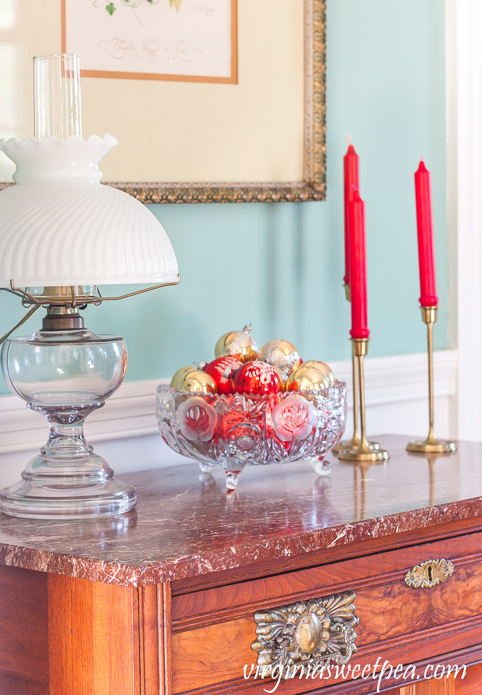 Decorations on a chest for Christmas including a trio of red candles in brass candle holders and an antique glass bowl filled with vintage red and gold Christmas ornament.