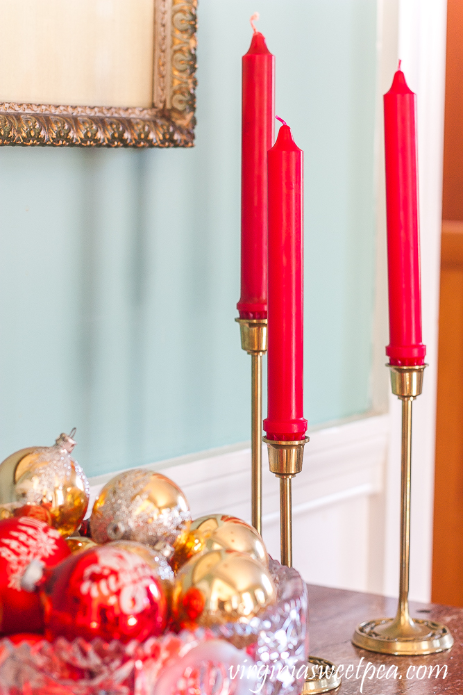 A trio of brass candle holders with a glass bowl filled with vintage red and gold Christmas ornaments in a dining room decorated for Christmas.