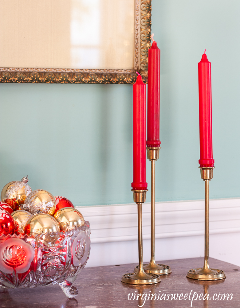 A trio of brass candle holders with a glass bowl filled with vintage red and gold Christmas ornaments in a dining room decorated for Christmas.