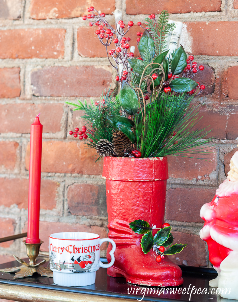 Large vintage Santa boot with greenery, vintage Christmas mug, brass holly candle holder with red candle.