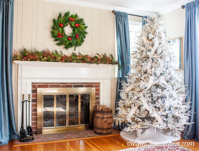 Christmas mantel with Williamsburg wreath and lighted garland on the mantel with red berries, Magnolia, and pine cones and a flocked Christmas tree decorated with snowflakes