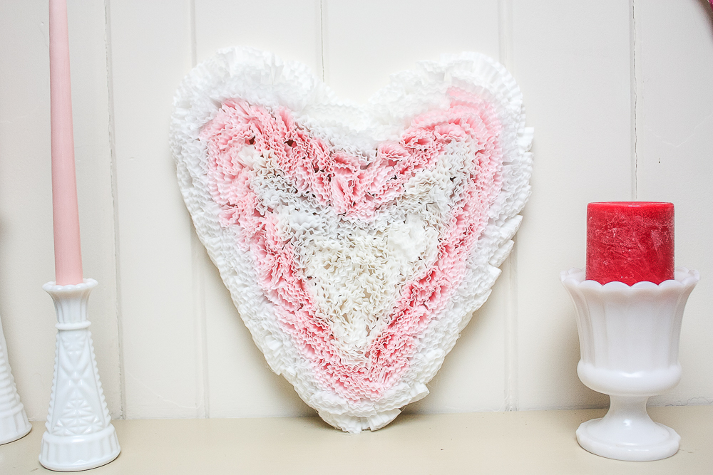 Cardboard heart decorated with pink and white cupcake liners on a mantel with white milk glass and pink and red candles