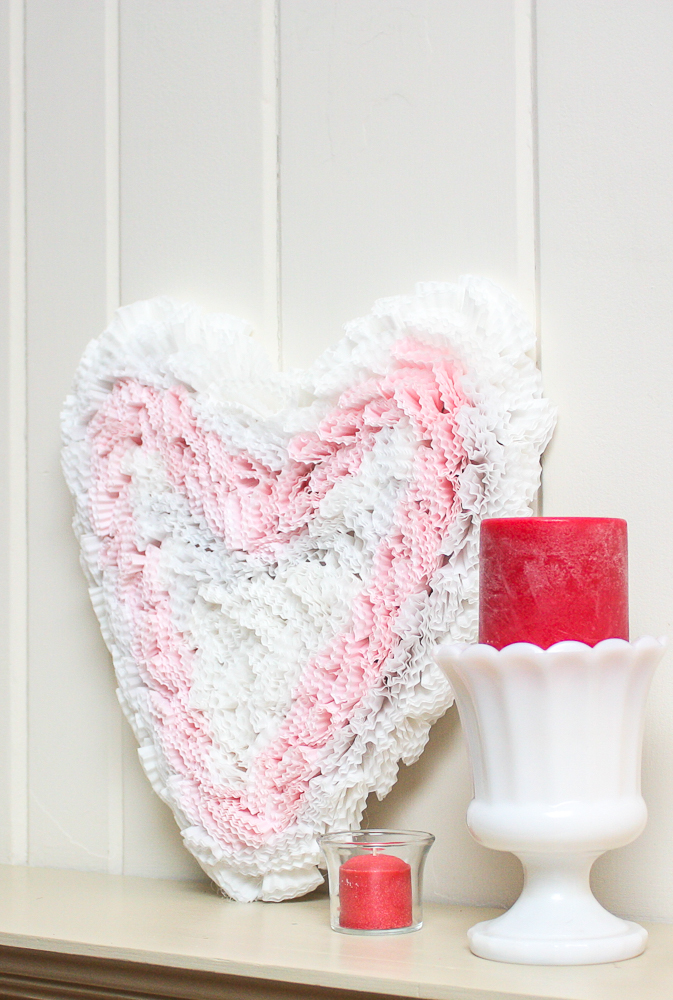 Handmade cupcake liner heart displayed on a mantel with dark pink candles in a clear glass votive holder and in a milk glass vase