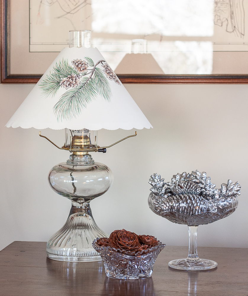 Deodar Cedar cones in a cut glass bowl with a glass compote filled with pine cones painted with silver paint beside an antique oil lamp with a paper shade with pinecones and greenery printed on the shade.