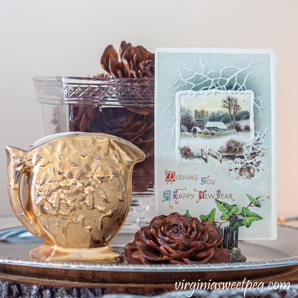 New Year vignette with an antique postcard, gold pitcher, and cones from a Deodar Cedar.