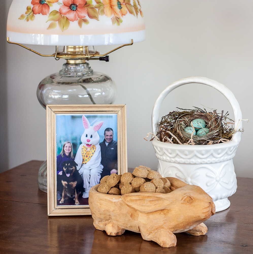 Easter vignette including a family picture with the easter rabbit, carved wood rabbit candy dish, McCoy basket with bird nest, and an oil lamp with a glass shade with flowers on it.