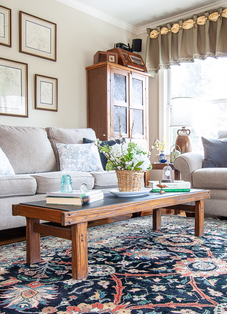 Coffee table decorated for spring with a flower arrangement in a basket, stacks of books, a blue antique insulator, carved bird, and an antique pocket watch under a glass cloche