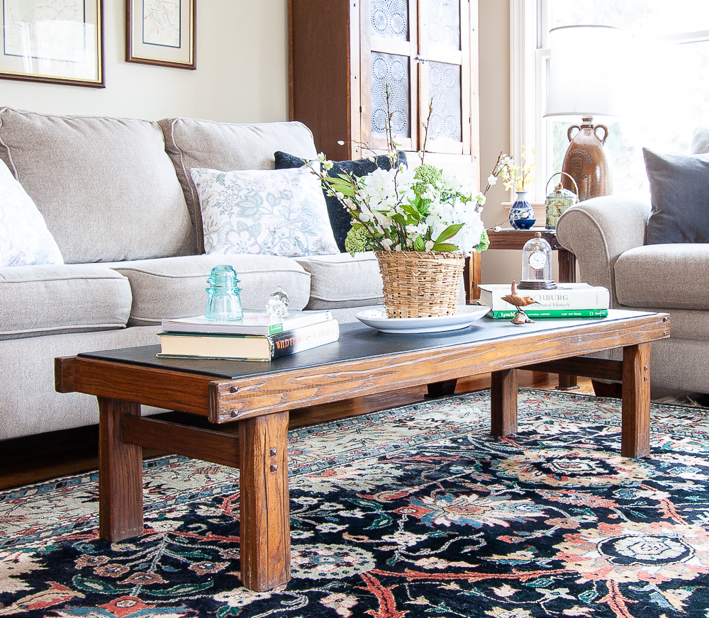 Coffee table decorated for spring with a flower arrangement in a basket, stacks of books, a blue antique insulator, carved bird, and an antique pocket watch under a glass cloche