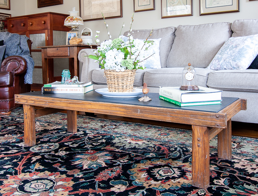 Coffee table decorated for spring with a flower arrangement in a basket, stacks of books, a blue antique insulator, carved bird, and an antique pocket watch under a glass cloche