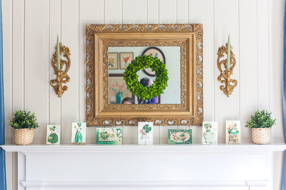 Mantel decorated for St. Patrick's Day with vintage St. Patrick's Day postcards, plants in woven baskets, a boxwood wreath, and green candles.