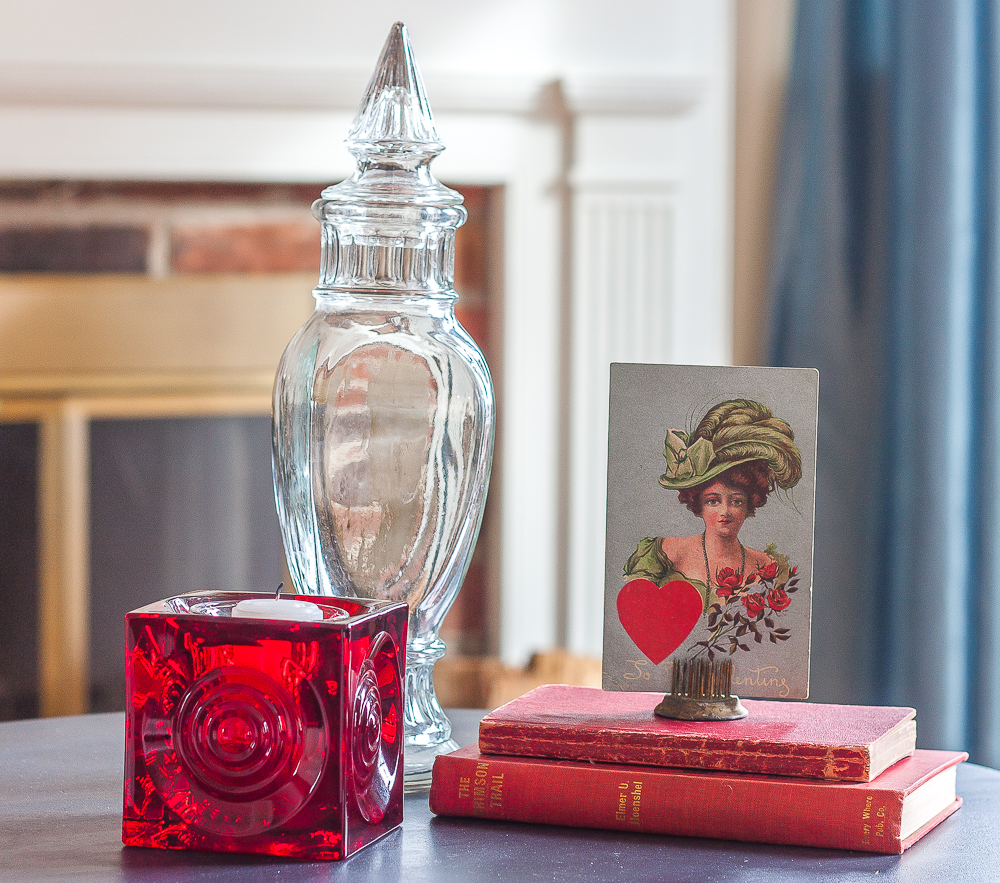 Valentine's Day vignette with a red Blenko candle holder, vintage red books, vintage early 1900s postcard, and a vintage glass candy jar