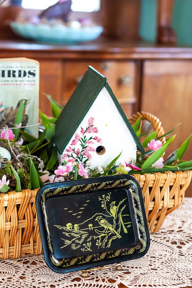 Small black tray with painted gold birds and scenery displayed in front of a spring centerpiece with a bird theme