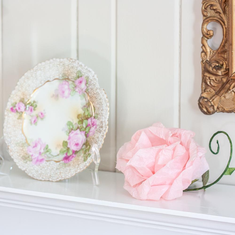 Pink crepe paper flower with a green stem and faux leaf and a plate with roses and gold pattern on the edge