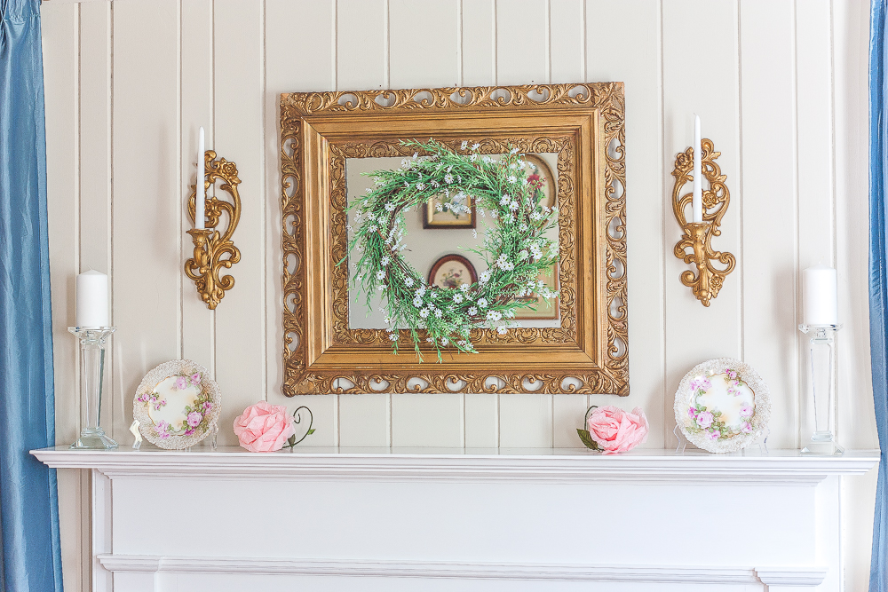 Gold mirror with a green and white wreath, gold sconces with white candles, floral plates, pink crepe paper flowers, and crystal candle holders with white candles.