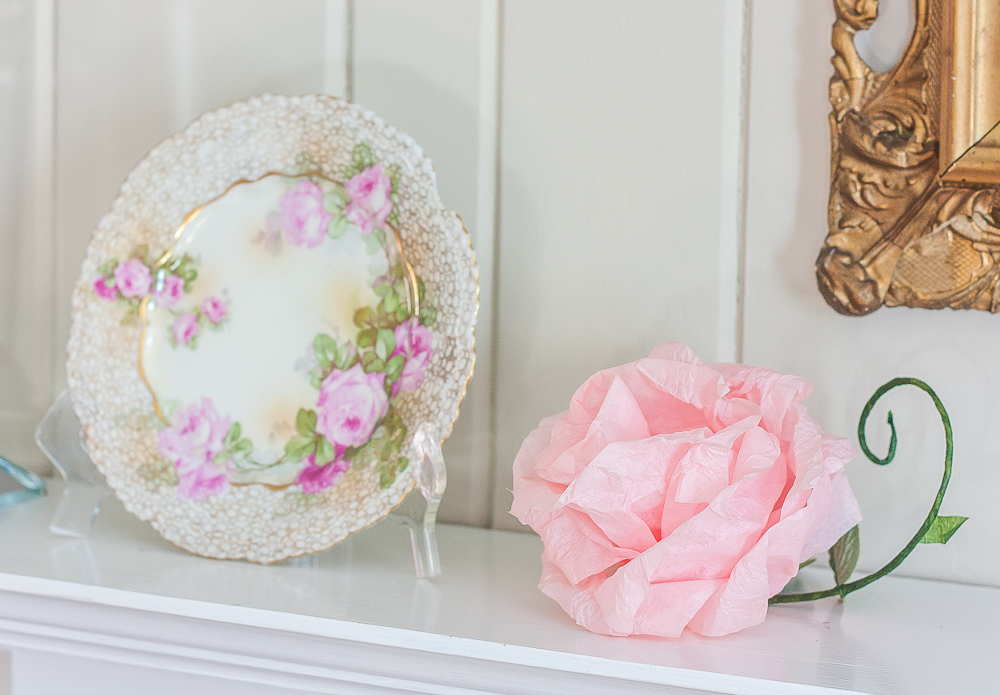 Pink crepe paper flower with a green stem and faux leaf and a plate with roses and gold pattern on the edge