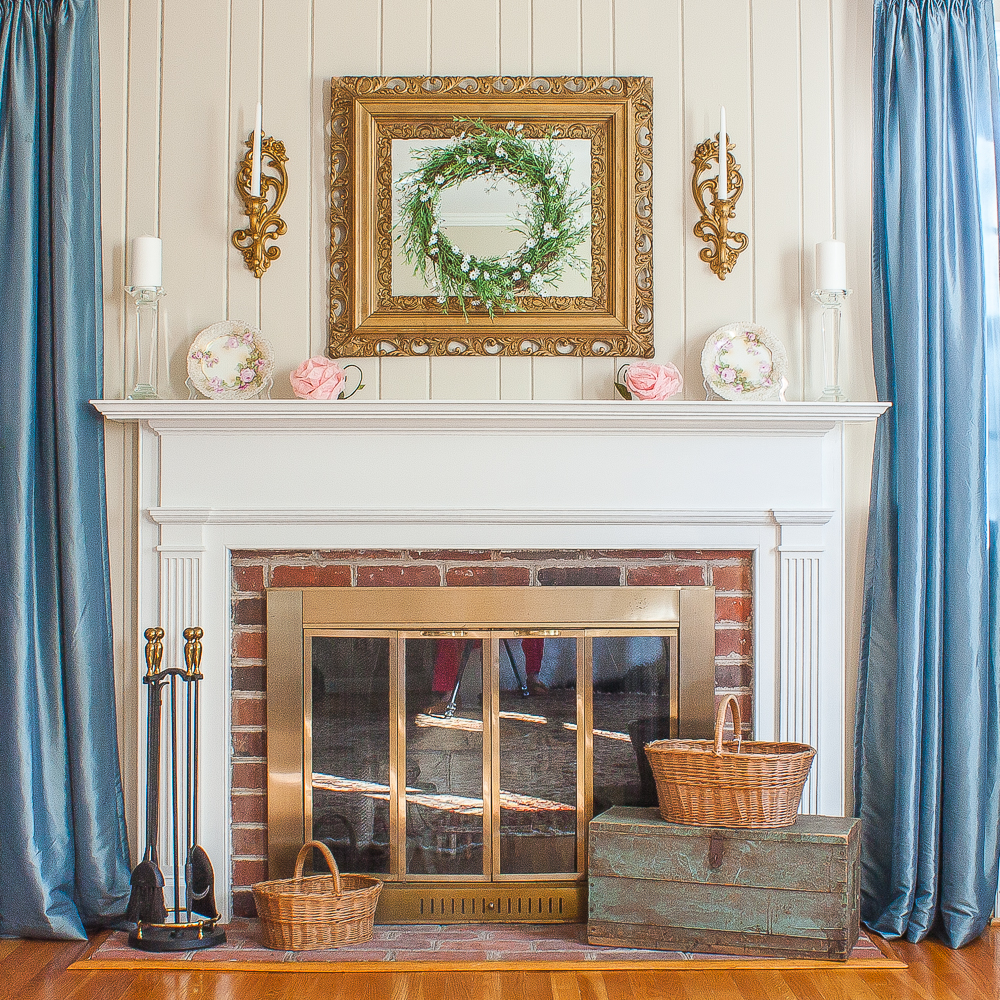 Mantel decorated for spring with pink flowers, a green wreath, white candles, and baskets on the fireplace hearth.