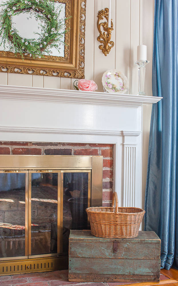 Mantel decorated for spring with pink flowers, a green wreath, white candles, and baskets on the fireplace hearth.