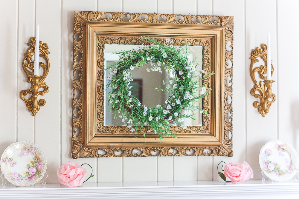 Gold mirror with a green and white wreath, gold sconces with white candles, floral plates, and pink crepe paper flowers.