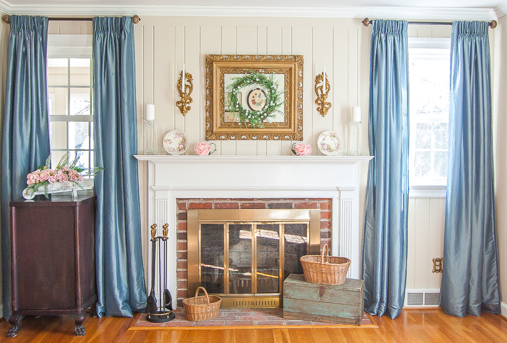 Mantel decorated for spring with pink flowers, a green wreath, white candles, and baskets on the fireplace hearth.