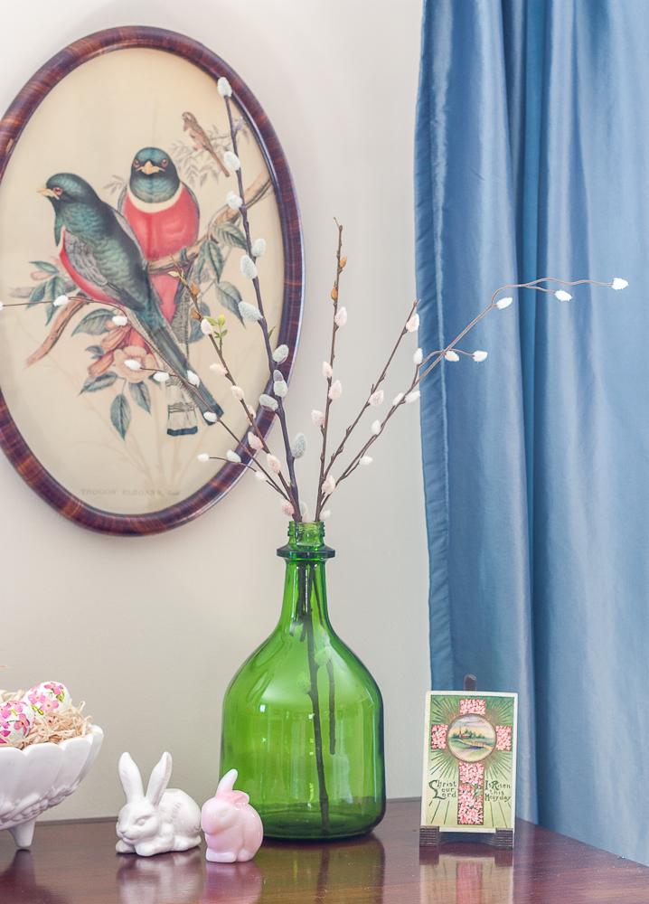 Easter vignette with a green bottle filled with pussy willow, an early 1900s Easter postcard, an Avon white rabbit candle holder, and a pink Fenton glass rabbit