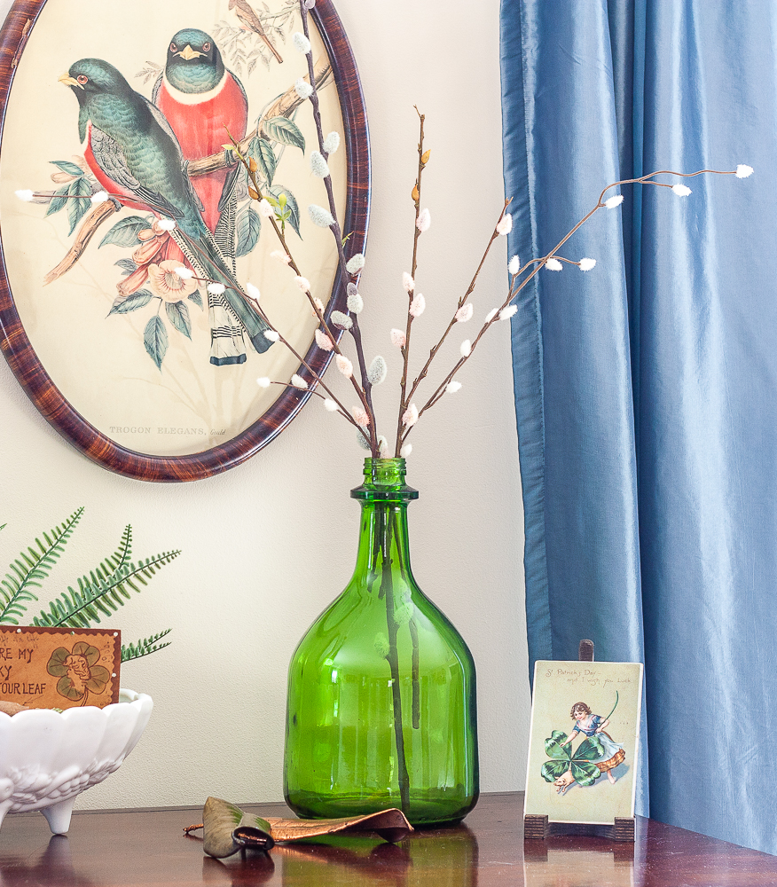 St. Patrick's Day vignette with a milk glass compote, green bottle with pussy willow, and a vintage St. Patrick's Day postcard