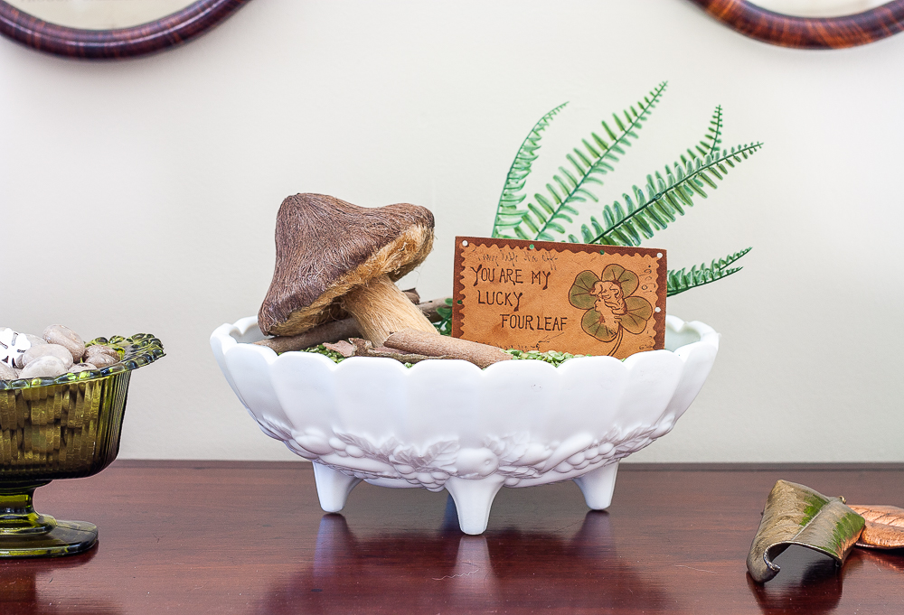 St. Patrick's Day vignette with a milk glass compote filled with split peas, ferns, a mushroom, and a vintage St. Patrick's Day postcard