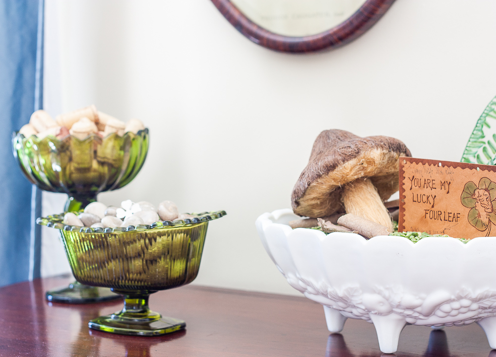 St. Patrick's Day vignette with a milk glass compote filled with split peas, ferns, a mushroom, and a vintage St. Patrick's Day postcard and two green glass compotes