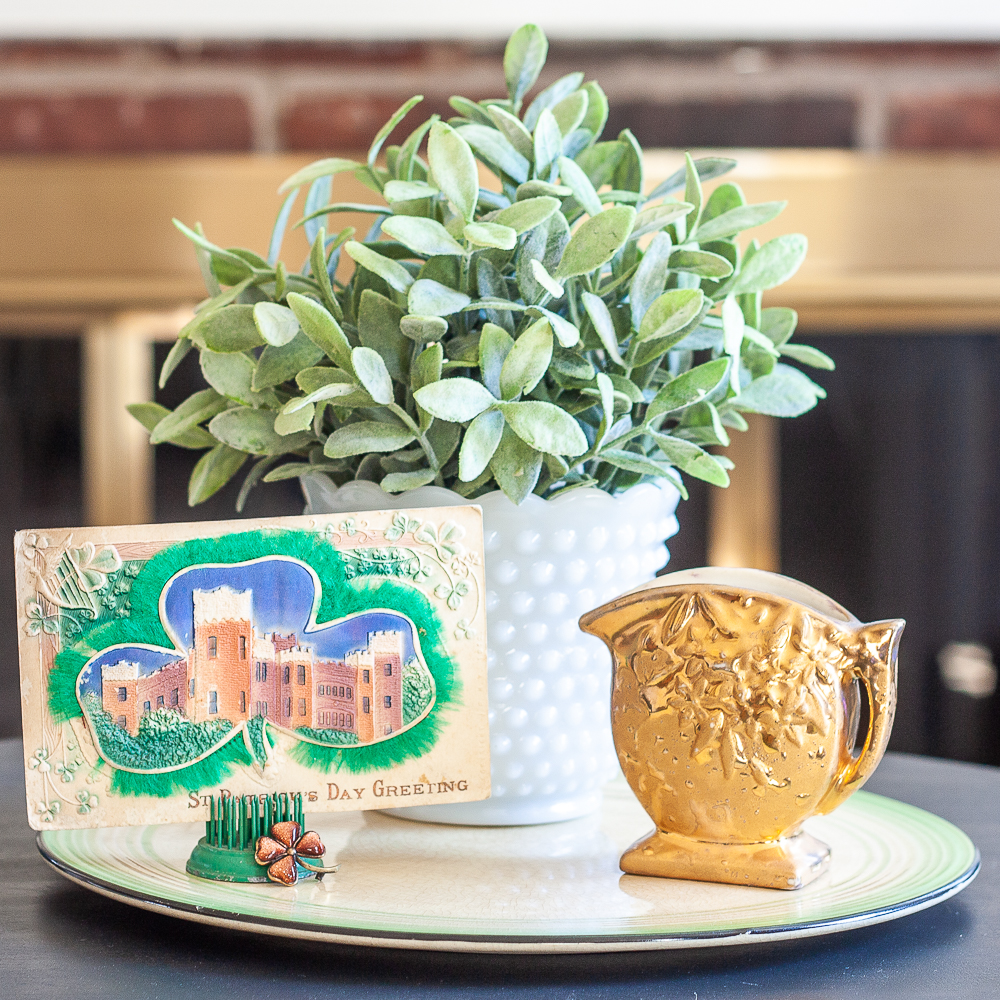 St. Patrick's Day vignette with a vintage postcard, gold pitcher, and a plant in a milk glass vase.