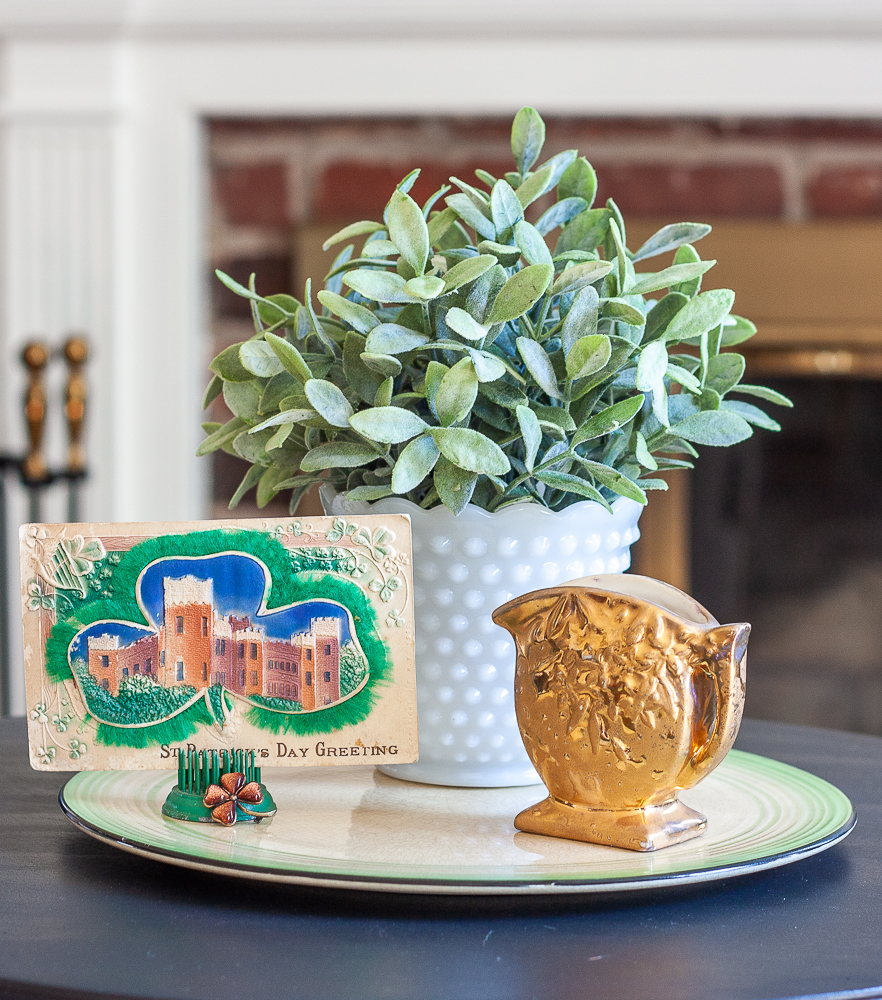 St. Patrick's Day vignette with a vintage postcard, gold pitcher, and a plant in a milk glass vase.