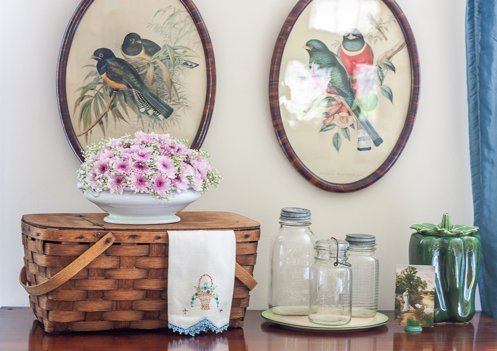 Vintage picnic basket with a floral arrangement, vintage embroidered tea towel, three vintage canning jars on a depression era round cake plate, McCoy green pepper cookie jar, and a 1908 postcard