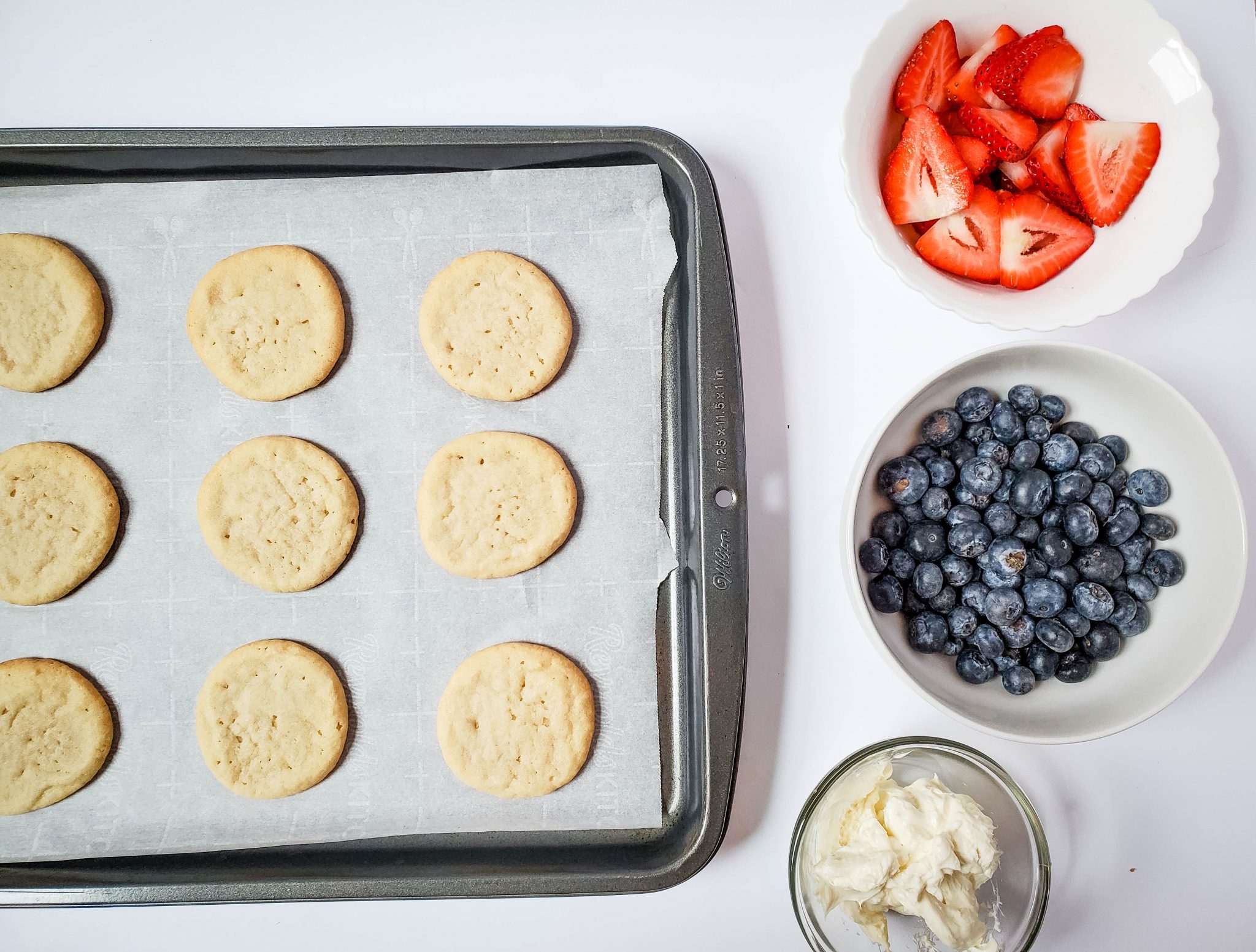 Sugar cookies on a parchment covered baking sheet, white bowl filled with strawberry slice, white bowl filled with blueberry slices, clear bowl filled with cream cheese frosting