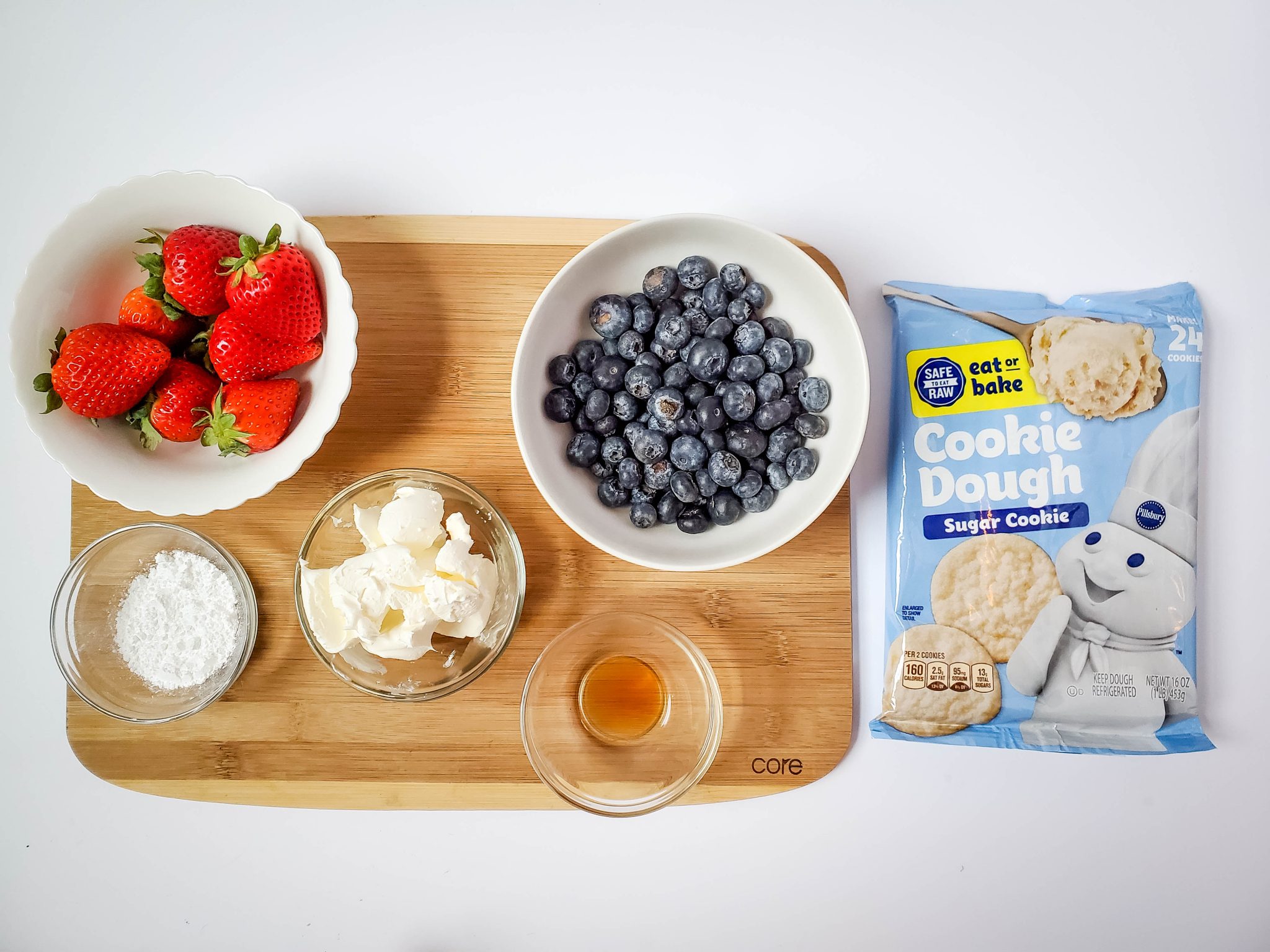 Pillsbury sugar cookie packaged dough, blueberries in a white bowl, strawberries in a white bowl, and clear glass bowls of powdered sugar, cream cheese, and vanilla