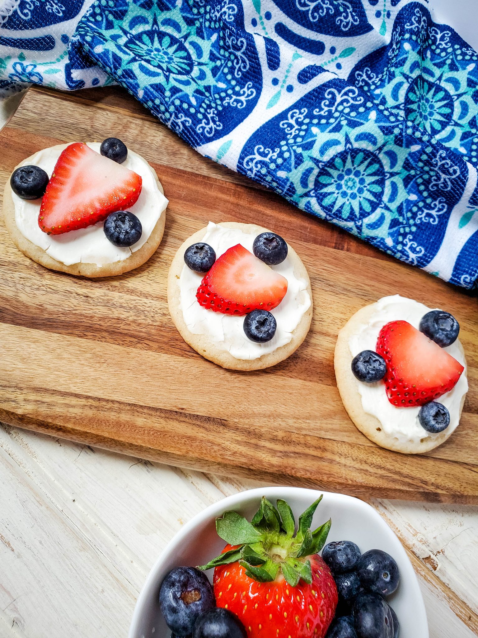 Sugar cookies decorated with a strawberry slice an three blueberries on a wood tray with a blue and white patterned tea towel in the background