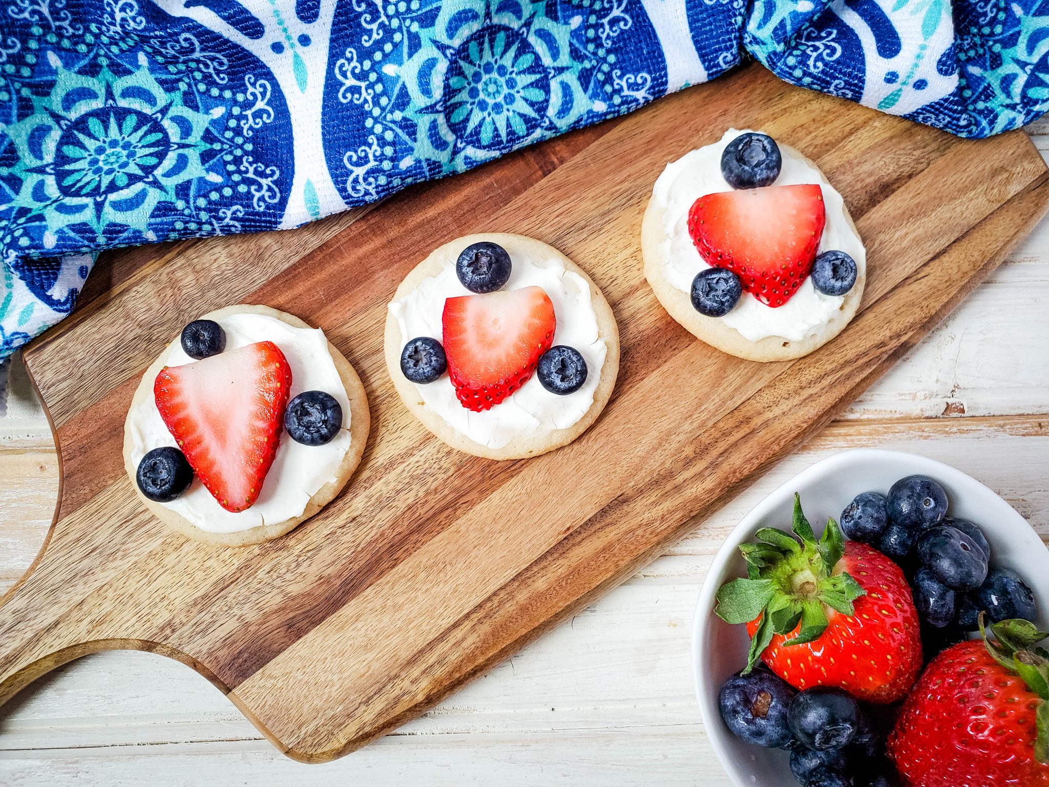 Three sugar cookies with white icing decorated with a strawberry slice and three blueberries on a wooden cutting board