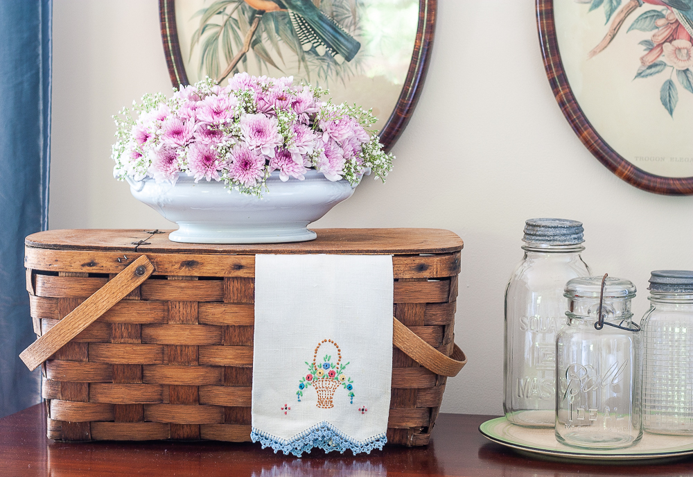 Vintage picnic basket with a floral arrangement, vintage tea towel, and three vintage canning jars on a depression era round cake plate