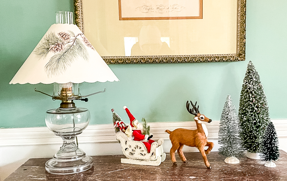 Santa in a sled with a reindeer pulling it, bottlebrush trees, and an antique lamp with a paper shade with white pine branches and cones printed on it