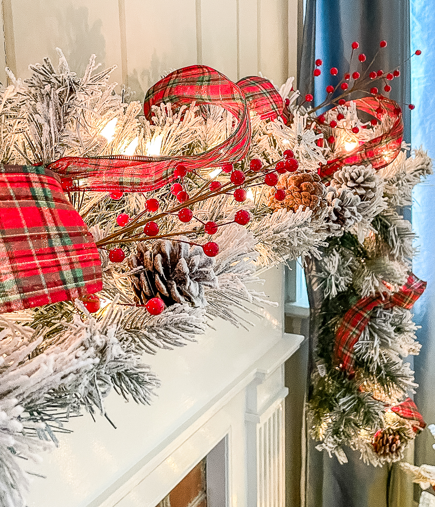 Fireplace decorated for Christmas with a flocked wreath and garland, red plaid ribbon, red berries, and red candles