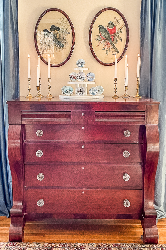 Antique chest decorated for winter with P. Buckley Moss ornaments on a cupcake stand and brass candle holders with white candles